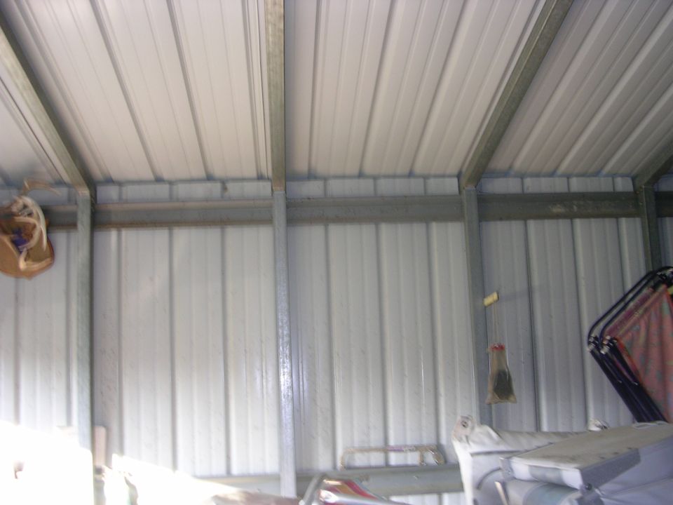 Inside a white metal carport with ribbed panels, galvanized posts and stored items