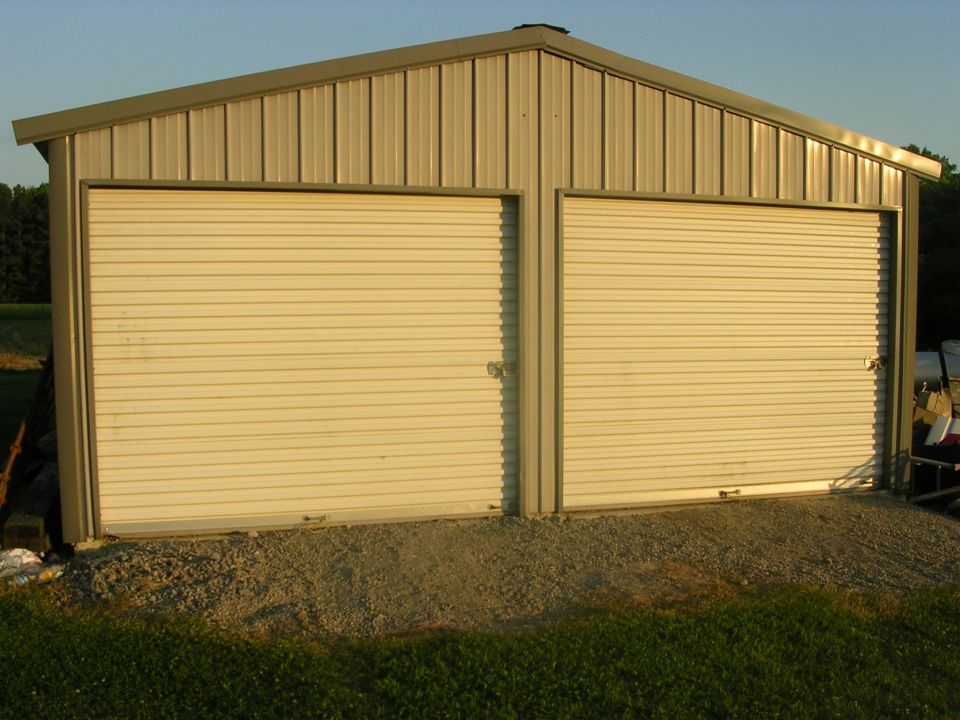 Tan metal garage building with two closed white roll-up doors on a gravel pad