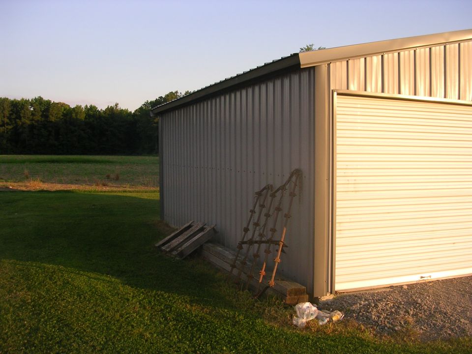 Side corner of a metal shop building with white roll-up door and leaning farm implement
