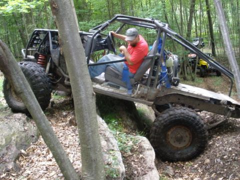 1995 Ford Bronco - A custom-built off-road buggy with a roll cage is driven over rocks in a wooded area.