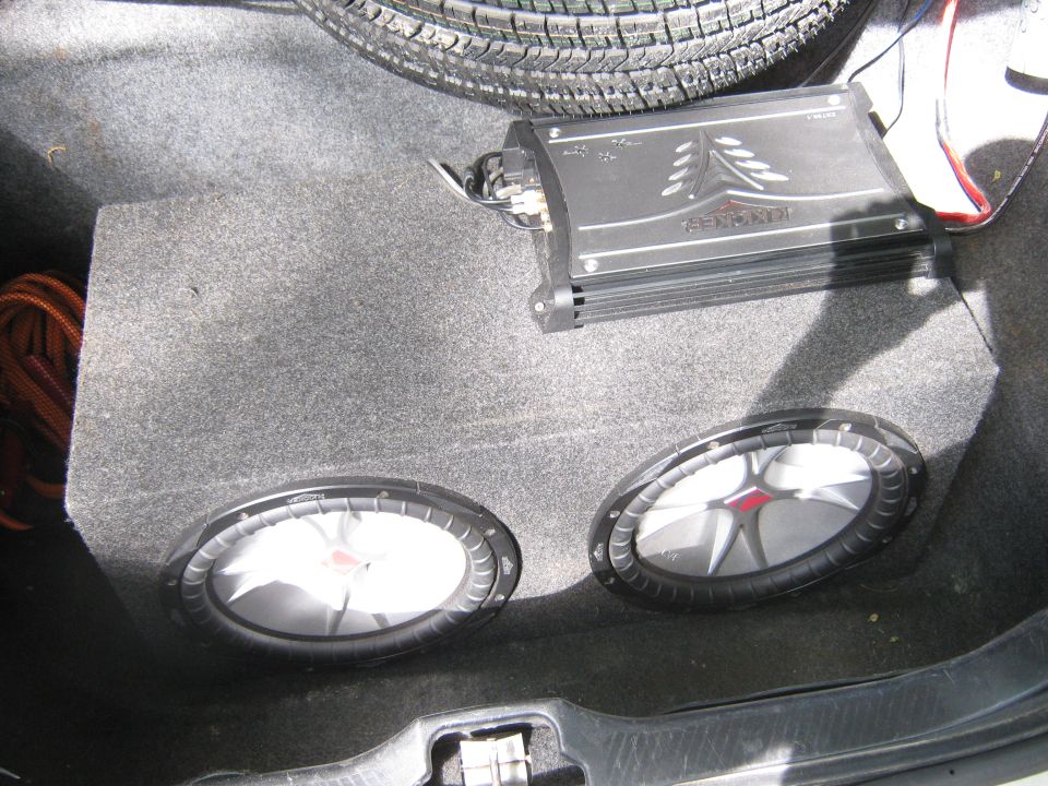 Car trunk interior showing two Kicker subwoofers in a box and a Kicker amplifier. Spare tire visible