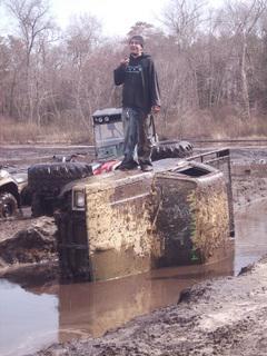 2005 Ford Excursion - A person stands atop an overturned vehicle stuck in a muddy area, with other vehicles visible in the background.