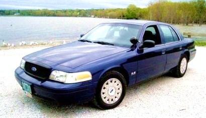 2003 Ford Crown Victoria - A blue 2003 Ford Crown Victoria is parked on a gravel surface near a body of water.