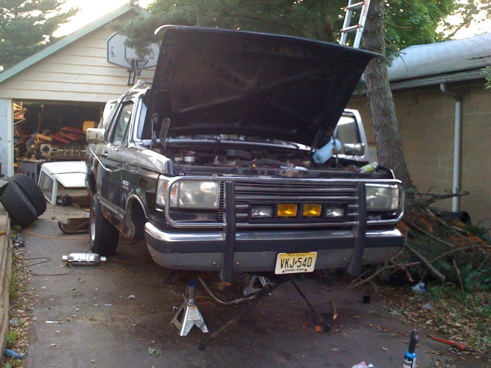 1987 Ford Bronco front view, hood open, on jack stands, in driveway with garage in background