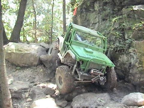 1992 Ford Bronco - Green tube-caged rock crawler on oversized off-road tires climbing a boulder pile on a wooded trail, front three-quarter view with a winch…