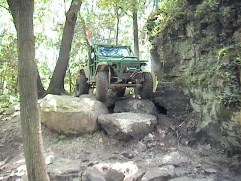 1992 Ford Bronco - A green off-road SUV with a tube front bumper, oversized mud tires and a lifted suspension climbs over large boulders on a rocky, tree-line…