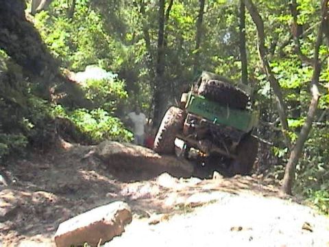 1992 Ford Bronco - An off-road vehicle with spare tires mounted on its roof navigates a challenging dirt path through a wooded area.