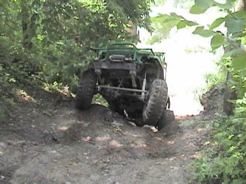 1992 Ford Bronco - A green tube-caged 4x4 buggy climbs over a steep dirt ledge on a wooded trail, front tires lifting and exposing the bare undercarriage, fro…