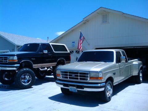 1994 Ford Bronco - A lifted black Bronco and a tan dually pickup truck are parked in a driveway on a sunny day.