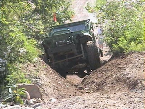 1992 Ford Bronco - A green open-top trail rig with an exposed roll cage twists across a deep dirt ditch, the front driver's tire hanging in the air while the…
