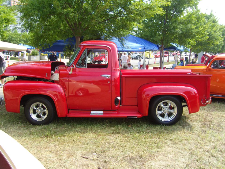 Red 1950s pickup truck with hood open, parked on grass next to an orange car