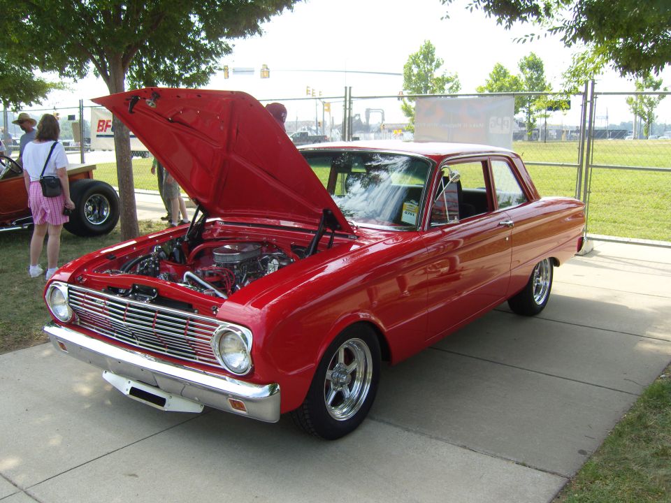 Red classic car, possibly a 1963 Ford Falcon, parked on pavement with hood open showing engine. Another car and people are visible in the background