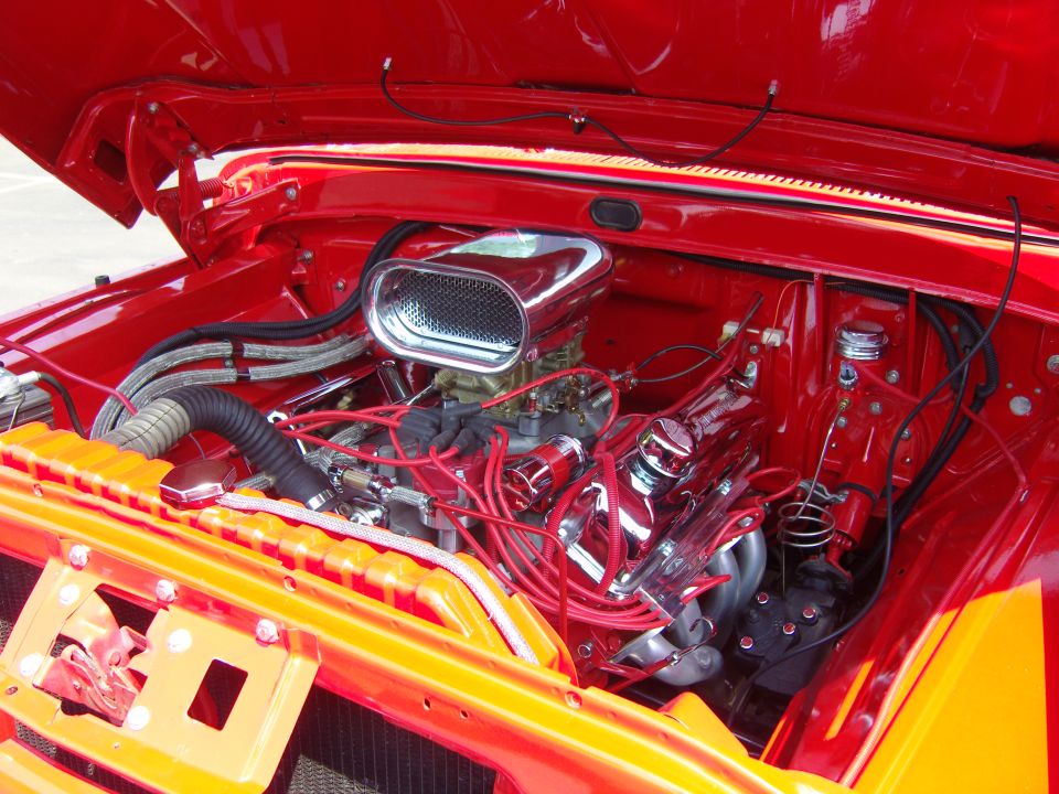 Engine bay of a classic car, showing a V8 engine with chrome valve covers, air cleaner, and red spark plug wires