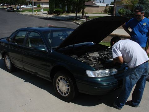 1993 Ford Taurus - Two men examine the engine of a dark green sedan parked on a suburban street.