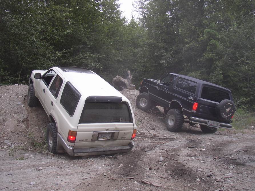 Lifted dark Ford Bronco on off-road dirt trail beside a white Toyota SUV in the woods.