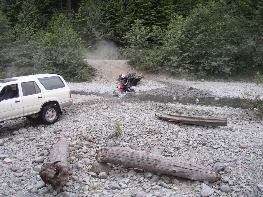 White SUV on rocky creek bed while a rider on a red ATV crosses a shallow stream near a forest trail