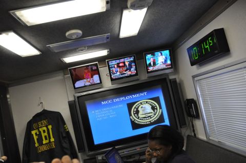 1996 Ford Bronco - Inside an FBI command center, multiple screens display news and presentations, with an FBI Commander jacket hanging nearby.