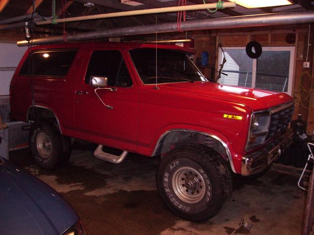 Red lifted Ford Bronco with white-letter Cooper tires parked in a garage, passenger-side front view