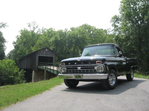 1966 Ford F-100 - Glossy black mid-1960s Ford F-100 styleside pickup photographed head-on from a low angle on a paved lane in front of a wooden covered bridg…