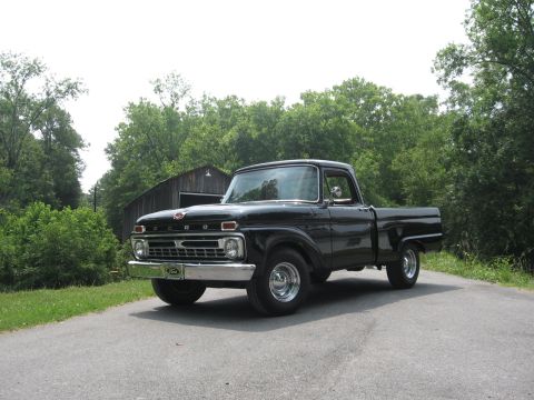 1966 Ford F-100 - Driver-side front three-quarter view of a gloss-black Ford styleside pickup parked on a paved country road with a weathered barn and green…