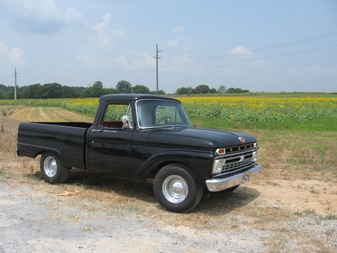 1966 Ford F-100 - A classic black Ford pickup truck is parked on a gravel road, with a field of sunflowers stretching out behind it under a partly cloudy sky.