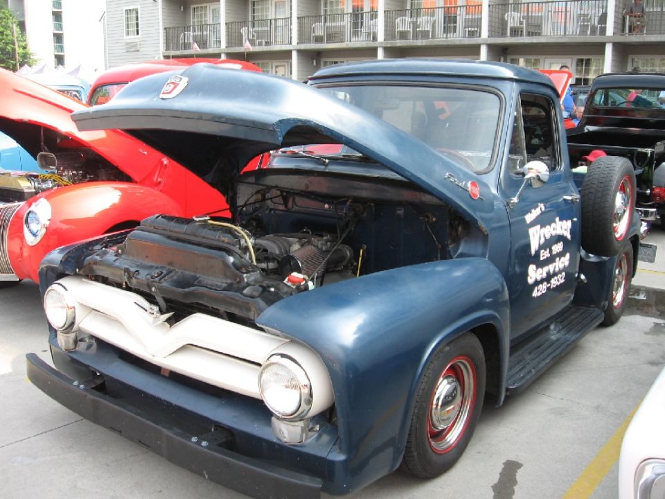 Blue Ford F-series truck with hood open, showing engine. "Wrecker Service" text on door. Red car behind it