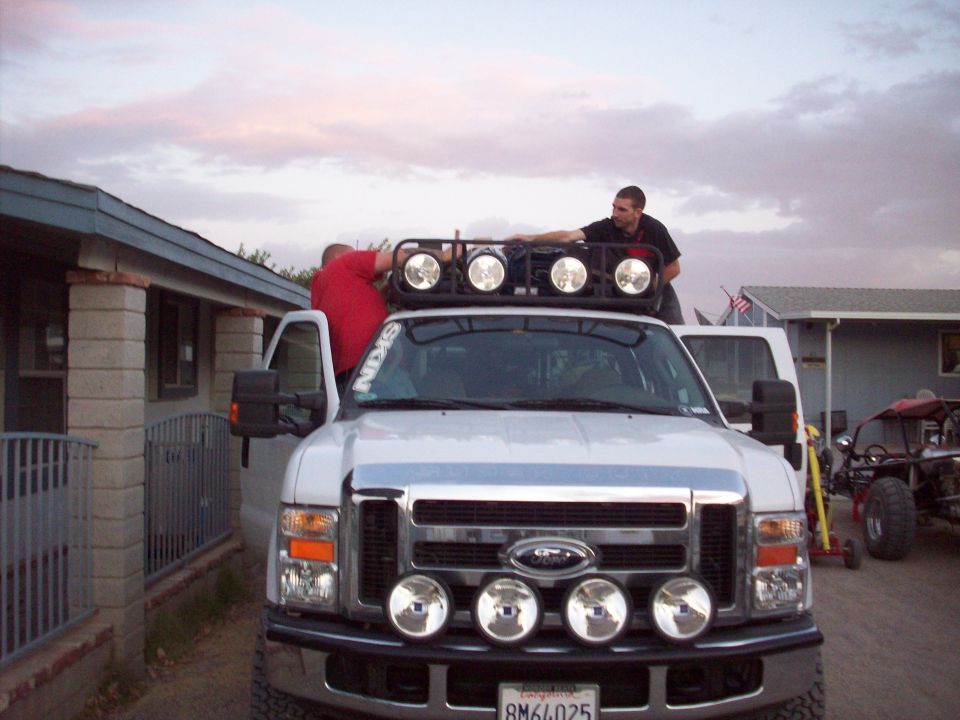 Front view of a white Ford Super Duty truck with a roof-mounted light bar and bumper lights. Two people are on or near the roof