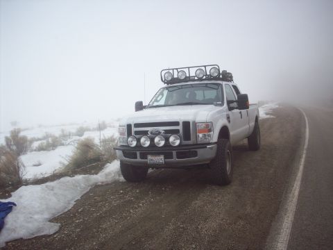 1995 Ford Bronco - A white Ford truck with auxiliary lights is parked on a dirt road with snow on the side.