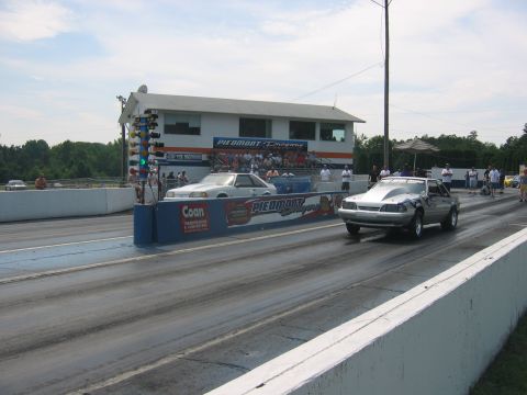 1992 Ford Mustang - Two cars race down a drag strip in front of a building with "Piedmont Dragway" signage.