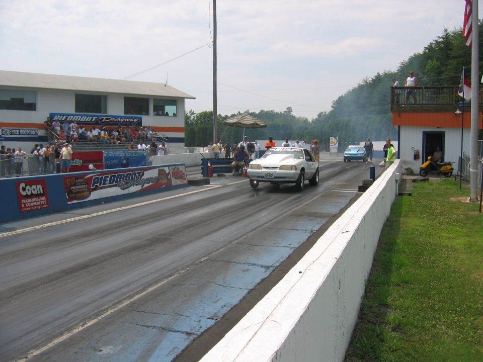 White 1992 Ford Mustang drag racing, with another car in the background. Spectators are in the stands