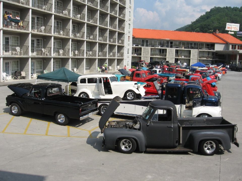 Several classic trucks parked in a lot, one dark gray truck in foreground with hood open