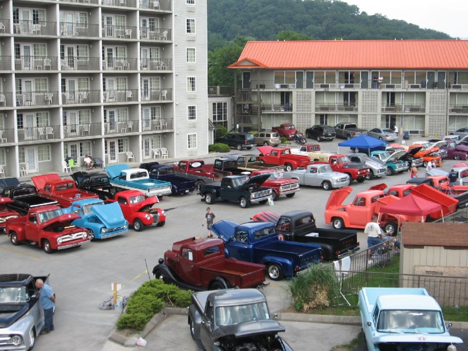Many pickup trucks parked in a lot outside a hotel. Some have hoods open