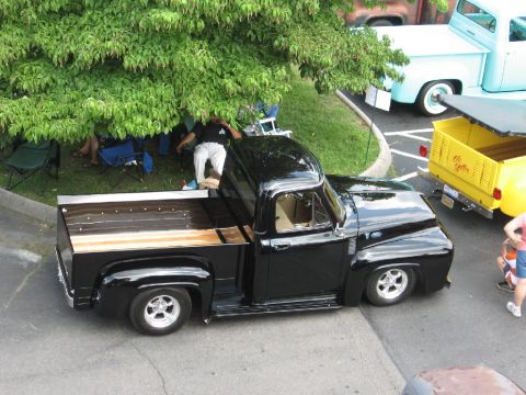 1966 Ford F-100 - A black pickup truck with a wooden bed is parked under a tree, with other vehicles nearby.