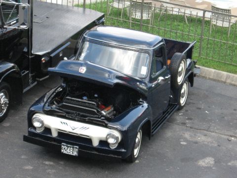 1966 Ford F-100 - This dark blue 1956 Ford F-100 pickup truck is displayed with its hood open, revealing the engine, and features a spare tire mounted on the…