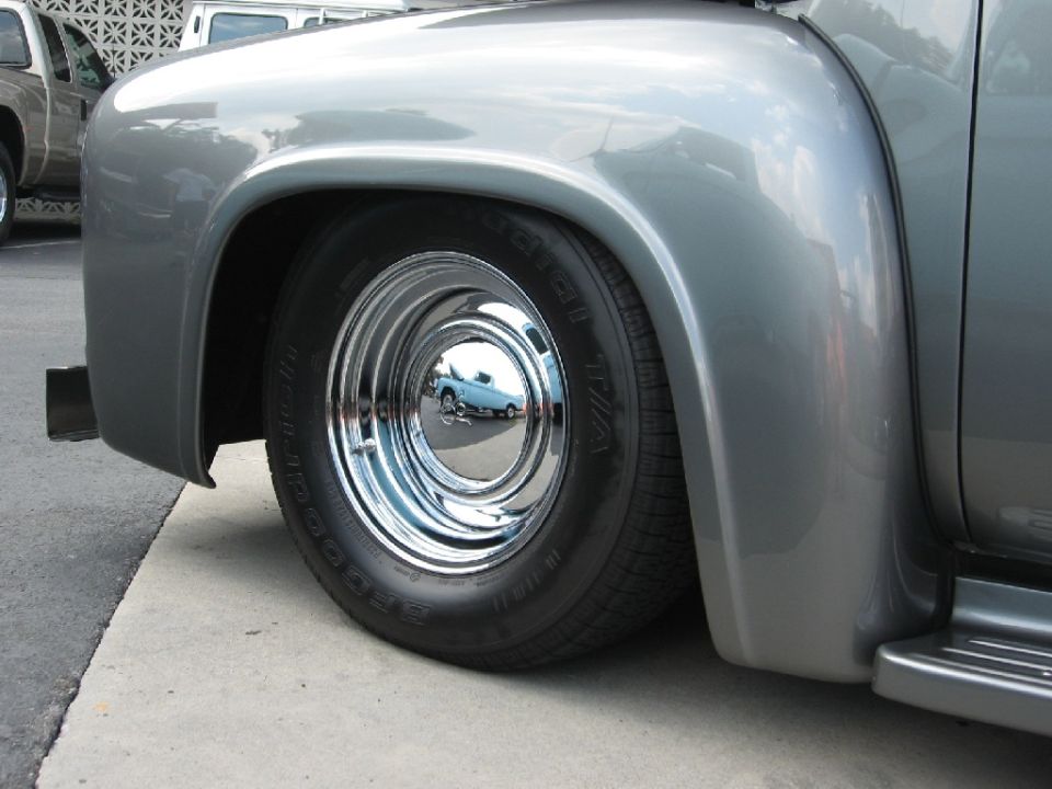 Close-up of a 1956 Ford F-series truck's front fender, chrome hubcap, and tire. Another truck is visible in the reflection