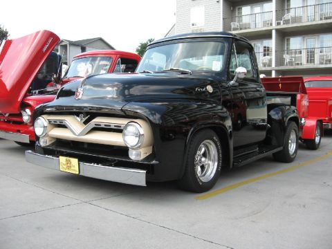 1966 Ford F-100 - A black 1956 Ford F-100 pickup truck is parked alongside two red trucks, one with its hood up.
