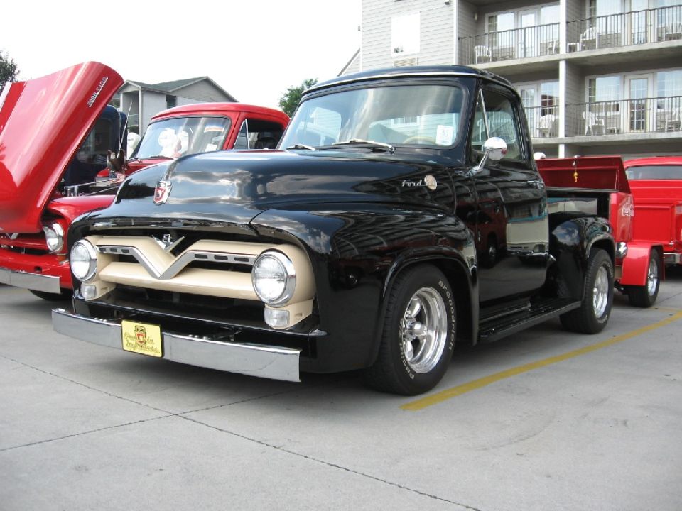 Black 1956 Ford F-100 pickup truck with tan grille, parked next to red trucks