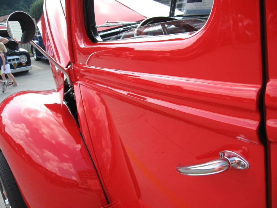 Close-up of a red vintage truck's door and fender. Another car is visible in the background