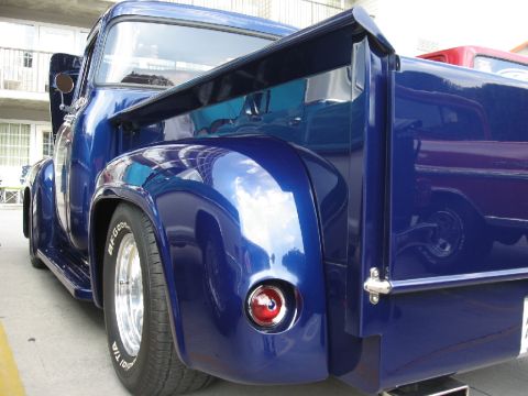 1966 Ford F-100 - A shiny blue pickup truck is seen from the rear quarter panel, a custom spoiler and chrome wheels. Another vehicle is reflected in the pain…