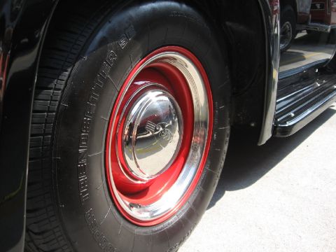1966 Ford F-100 - A close-up view of the wheel on a black 1966 Ford F-100, a red rim and a chrome hubcap. Another vehicle is visible in the background.