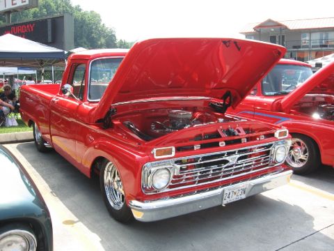 1966 Ford F-100 - A bright red 1966 Ford F-100 pickup truck is displayed with its hood open, revealing the engine bay, at an outdoor car show.