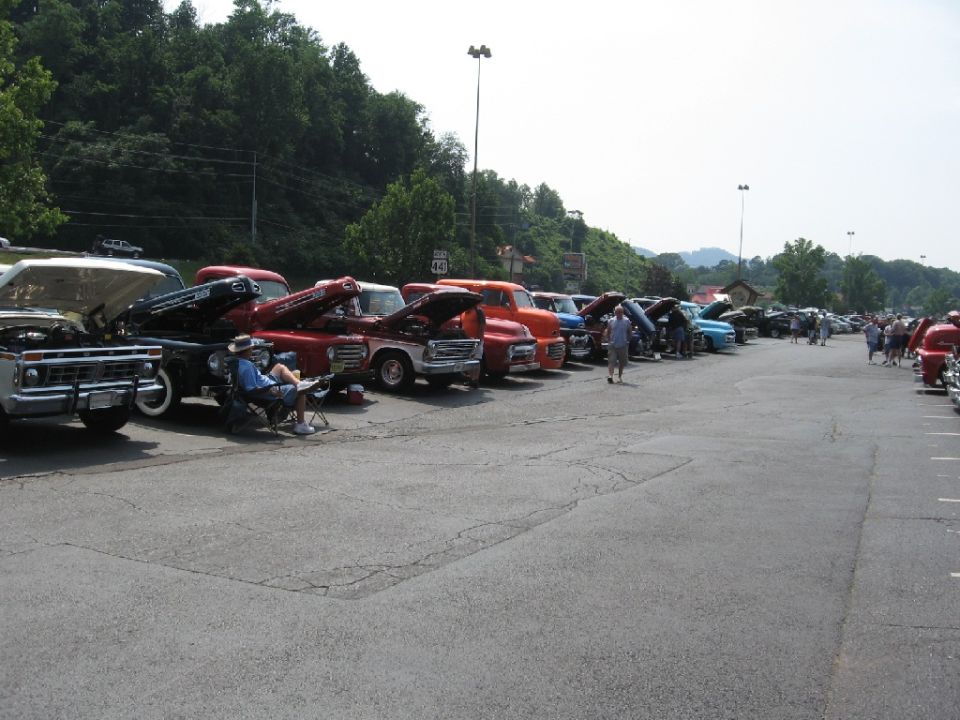Several classic pickup trucks parked in a lot, some with hoods up. A man sits in a chair near the front
