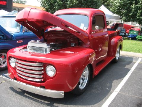 1966 Ford F-100 - A bright red pickup truck with its hood raised to reveal the engine, parked next to a blue truck at an outdoor event.