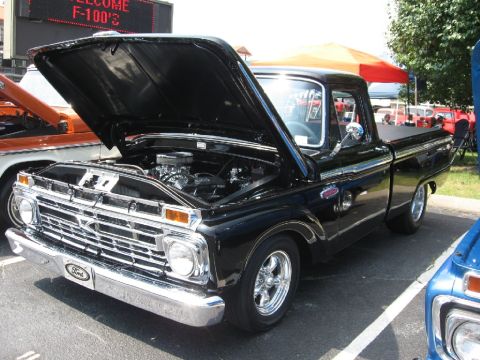 1966 Ford F-100 - A black Ford F-100 pickup shown from the front three-quarter angle with its hood raised, revealing a chrome-dressed V8 engine, and fitted w…