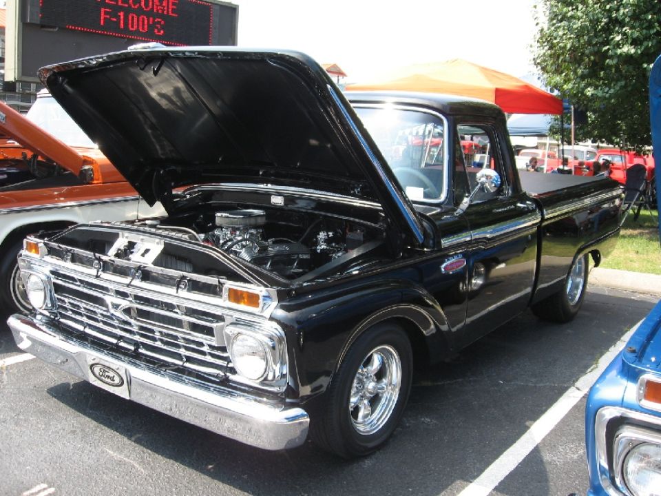 Black Ford F-100 pickup with hood up showing V8 engine at a car show