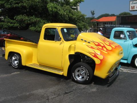 1966 Ford F-100 - A bright yellow pickup truck with custom flame paint on the hood and chrome wheels is parked next to a blue truck.