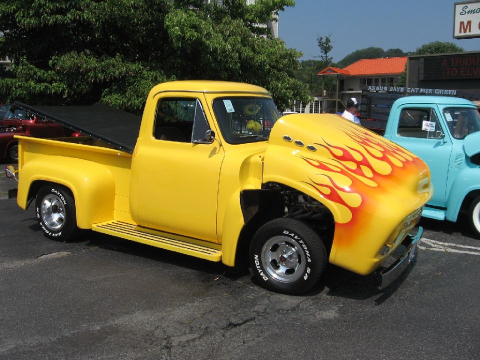 Yellow pickup truck with flame decals on hood, custom wheels, and black tonneau cover. Another blue truck is visible