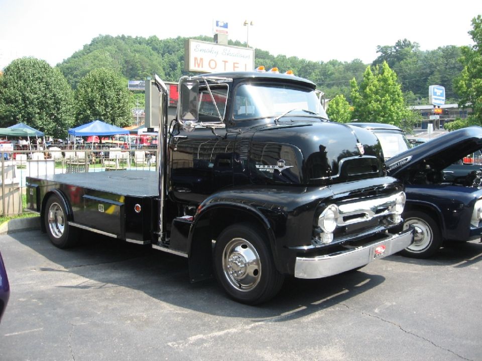 Black flatbed truck with chrome grille and dual rear wheels. Another truck with hood open is parked next to it
