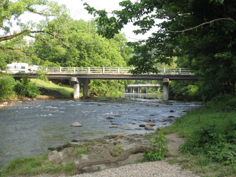 Two RVs drive across a bridge over a rocky river, with trees on both sides