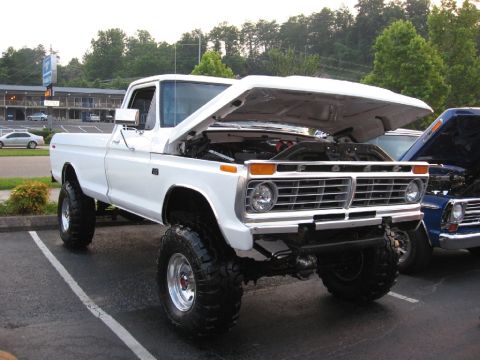 1966 Ford F-100 - White Ford F-Series pickup truck with hood open, lifted suspension, and large tires parked next to a blue truck.